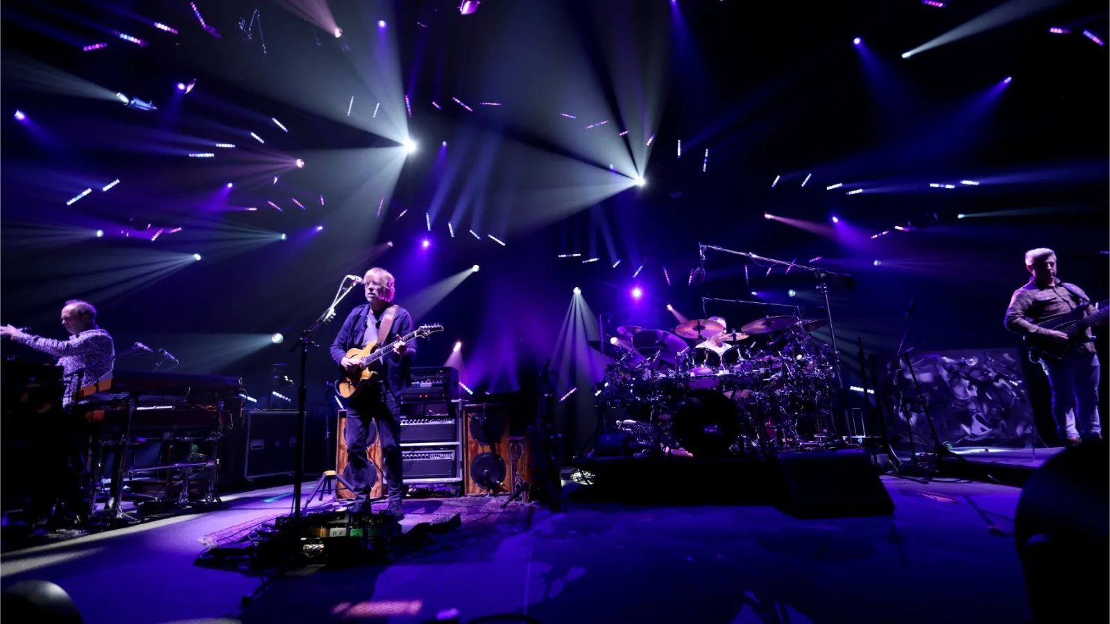 Multitud de fans de Phish bailando en un festival al aire libre con escenario iluminado de fondo